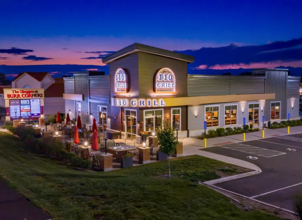 Aerial view of 110 Grill at The Shoppes at Burr Corners in Manchester, Connecticut, photographed during blue hour with exterior lights glowing.