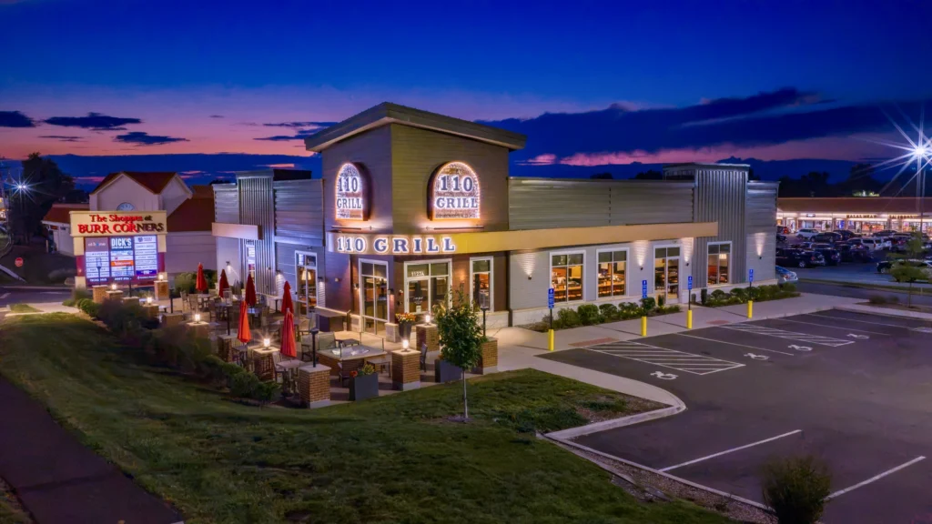 Blue-hour aerial view of 110 Grill at The Shoppes at Burr Corners in Manchester, Connecticut, with the restaurant illuminated.