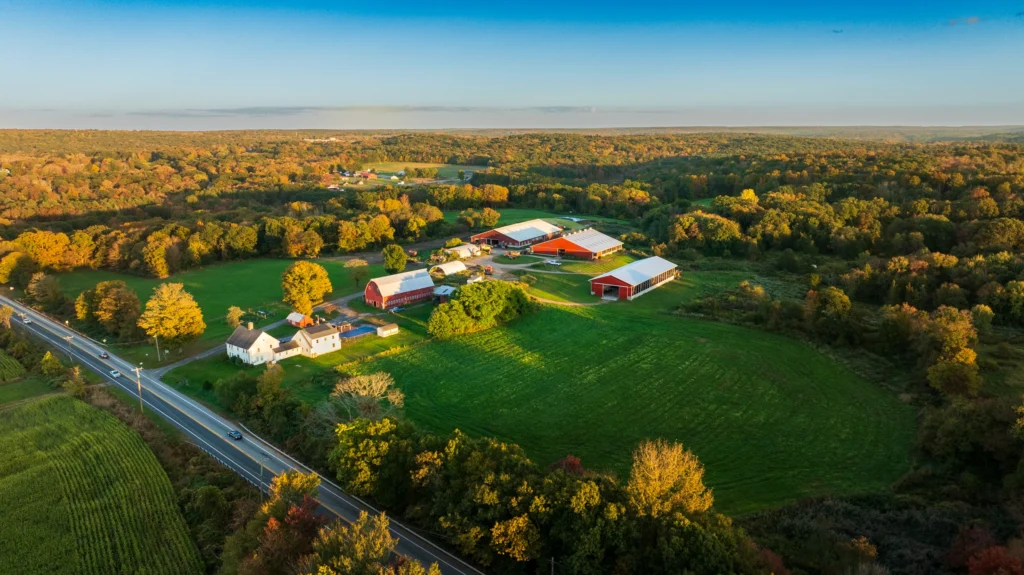 Wide aerial view of Hills Farm in Hebron, Connecticut, showing barns, open fields, a roadway and surrounding autumn woodland.