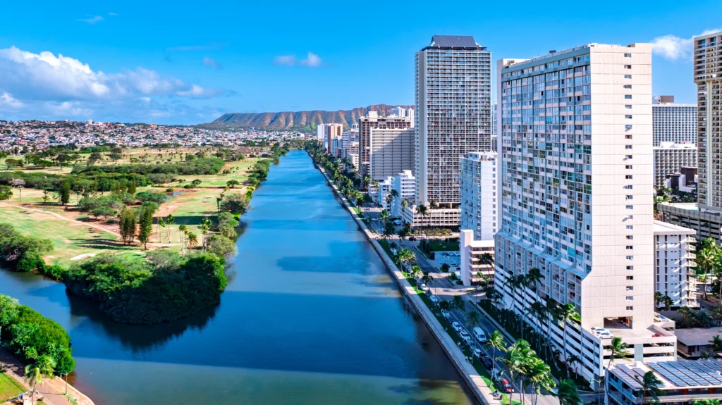 Aerial view over Honolulu, Hawaii, showing high-rise buildings, a canal and dense urban development extending toward the coast.