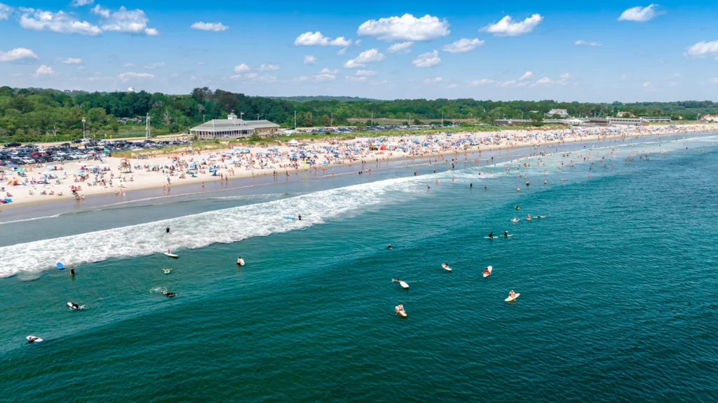Aerial view of Narragansett Beach in Rhode Island with surfers in the water, breaking waves and beachgoers along the shore.