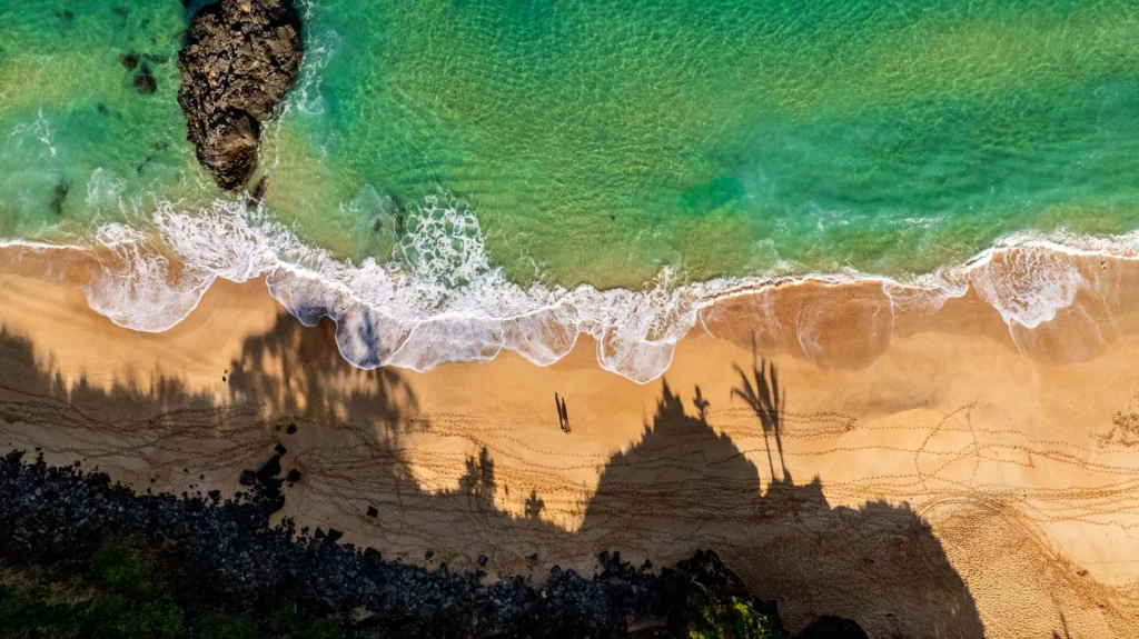 Top-down aerial photograph of a sandy Oahu beach with turquoise surf, breaking waves and dense coastal vegetation.