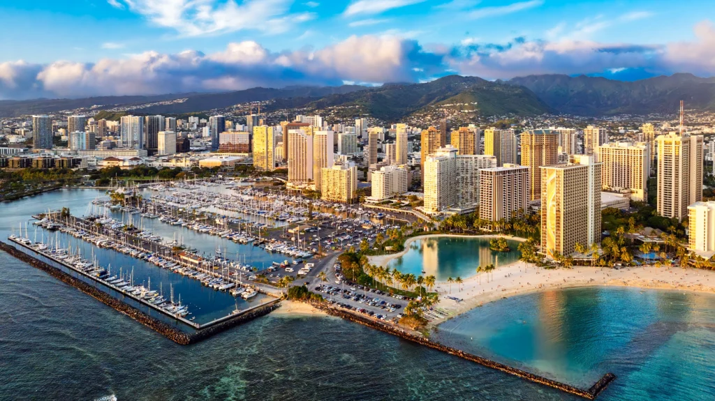 Wide aerial view of Waikiki on Oahu, Hawaii, showing the marina, beachfront high-rises, turquoise water and mountains in the distance.