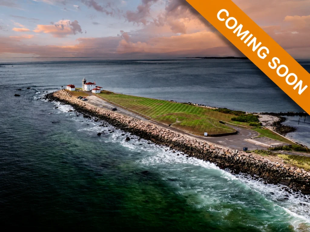 Aerial view of Watch Hill Lighthouse on a rocky point in Rhode Island at sunset, surrounded by ocean water.