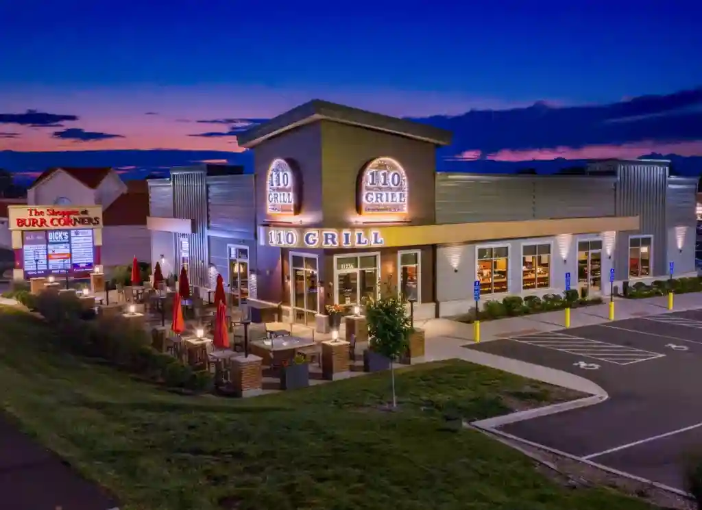 Blue-hour aerial view of 110 Grill at The Shoppes at Burr Corners in Manchester, Connecticut, with the restaurant illuminated.